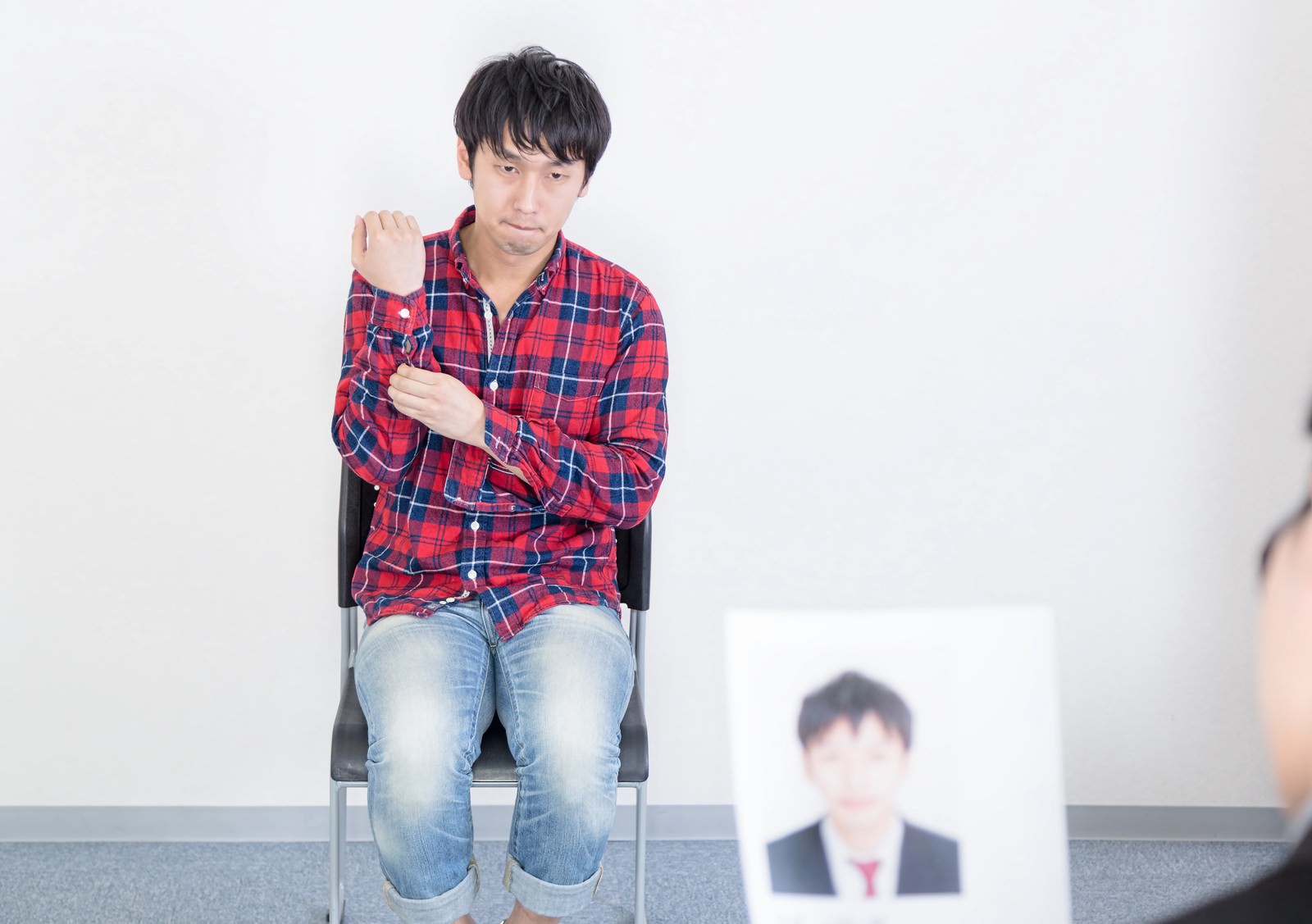 赤紺チェック柄シャツの男性が椅子に座り、肩をすくめながら両腕を上げている。背景奥にぼやけた証明写真が見える［モデル：大川竜弥］