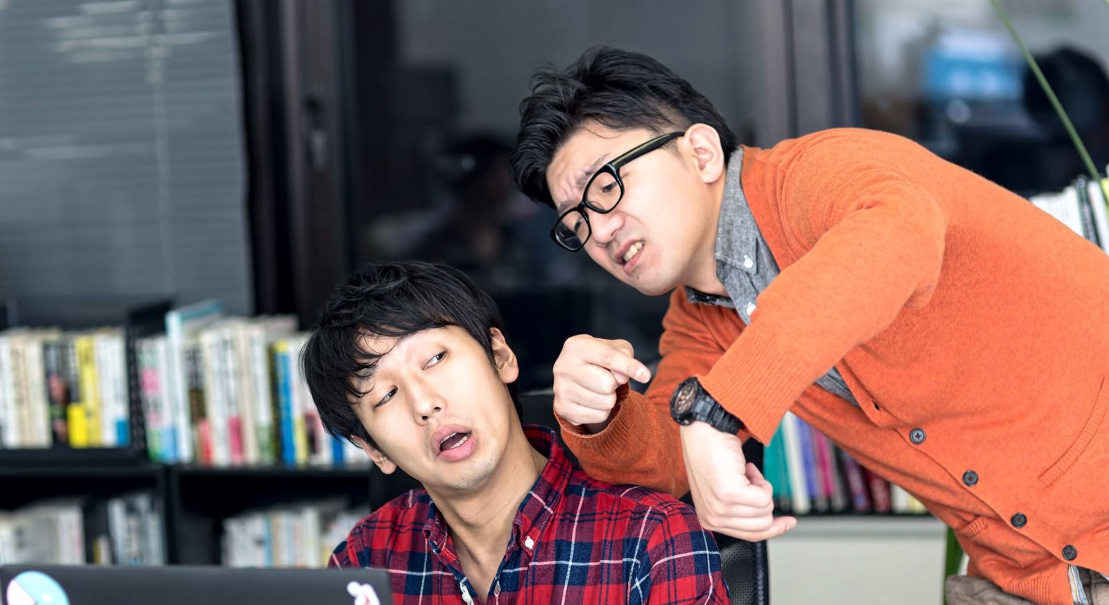 オレンジのカーディガン姿の眼鏡男性が、赤チェックシャツの男性に業務指示する様子［モデル：大川竜弥 OZPA］