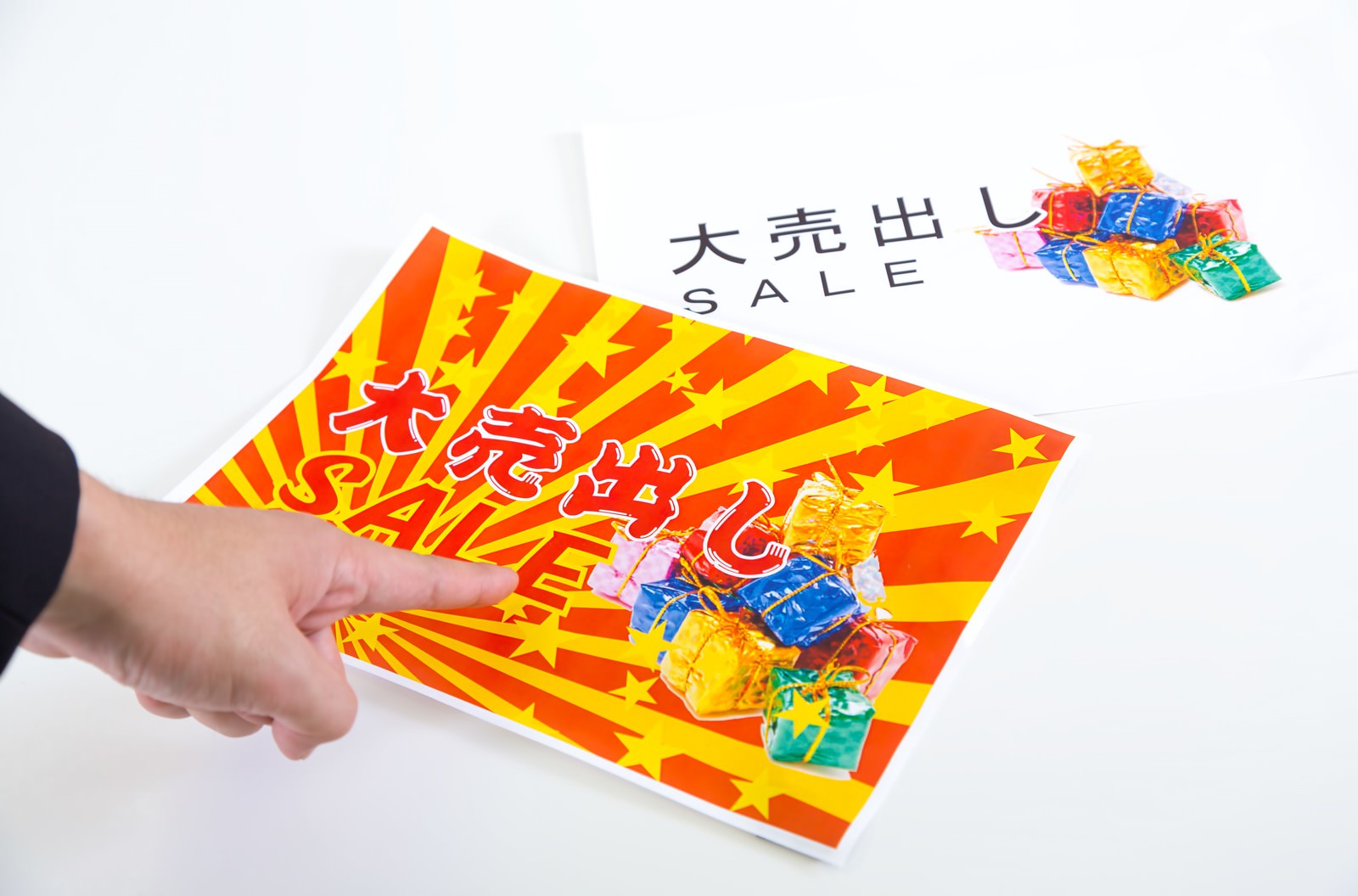 赤黄色の放射線パターンが入った大売出しチラシを左手で保持し、カラフルなプレゼントボックスが積み重なる写真