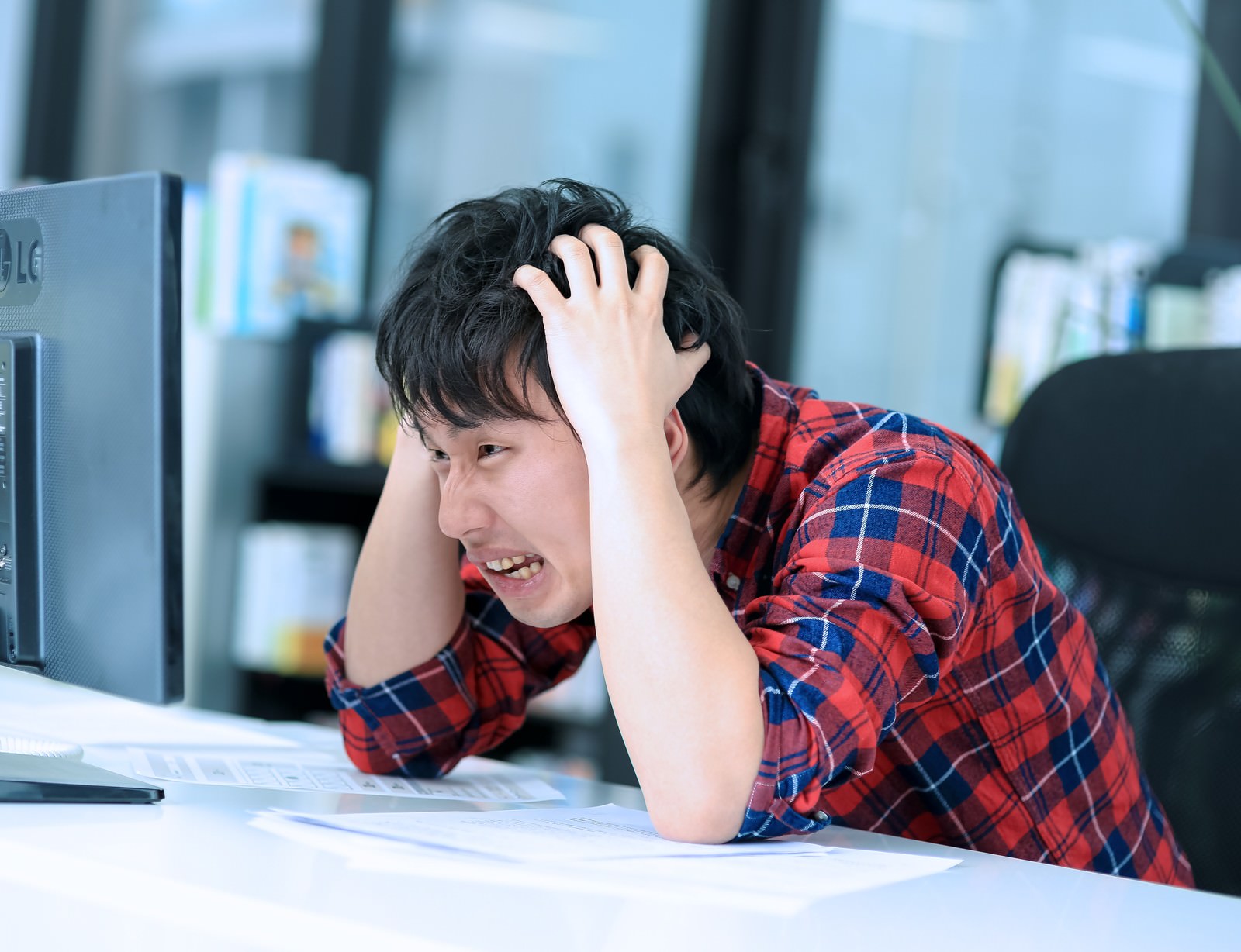 納品直後の校正ミスに気づき両手で頭を抱えて絶望するチェックシャツの男性［モデル：大川竜弥］