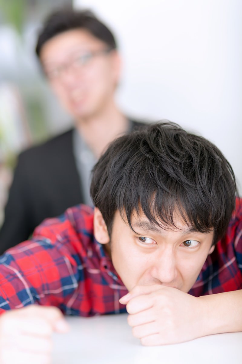 机に伏せて上目遣いで睨むチェックシャツの男性と背後にぼやけるスーツ姿の男性［モデル：大川竜弥］