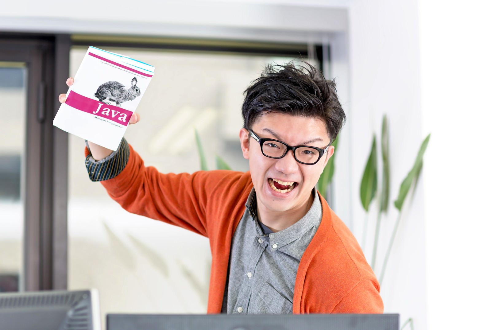 オレンジのカーディガンを着た眼鏡の男性が、パソコンの前でピンク色のカードを掲げて笑っている［モデル：OZPA］