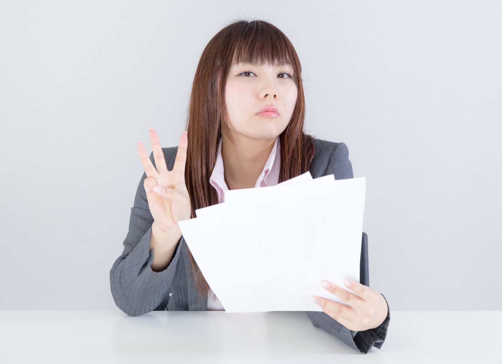 グレーのスーツ姿の女性が白い資料を両手で持ち、左手で3本指を立てながら上方を見つめている［モデル：Lala］