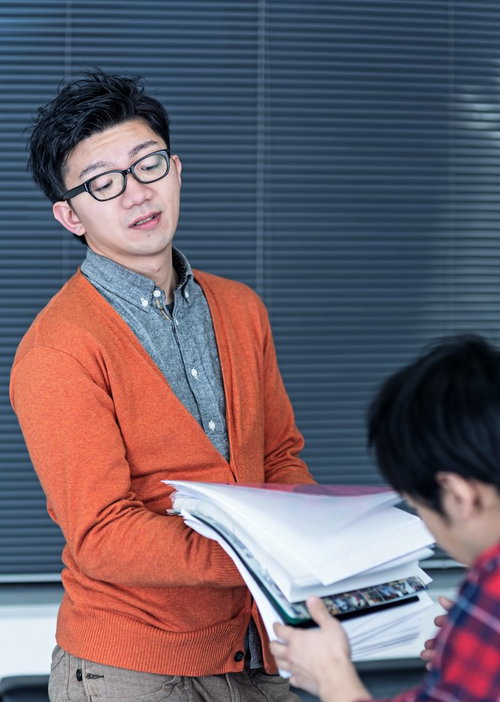白い書類の束を両手で持つ眼鏡の男性