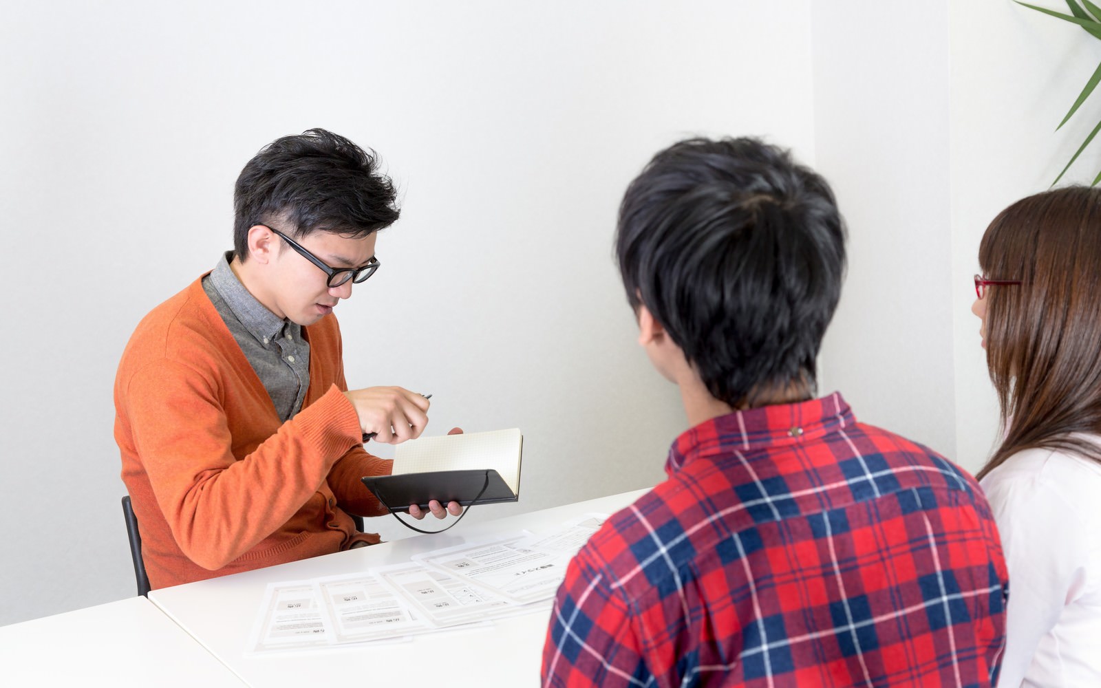 オレンジニットの男性がタブレットを持ち、赤チェックシャツ男性と眼鏡の女性と向き合って打ち合わせをしている［モデル：大川竜弥 Lala OZPA］