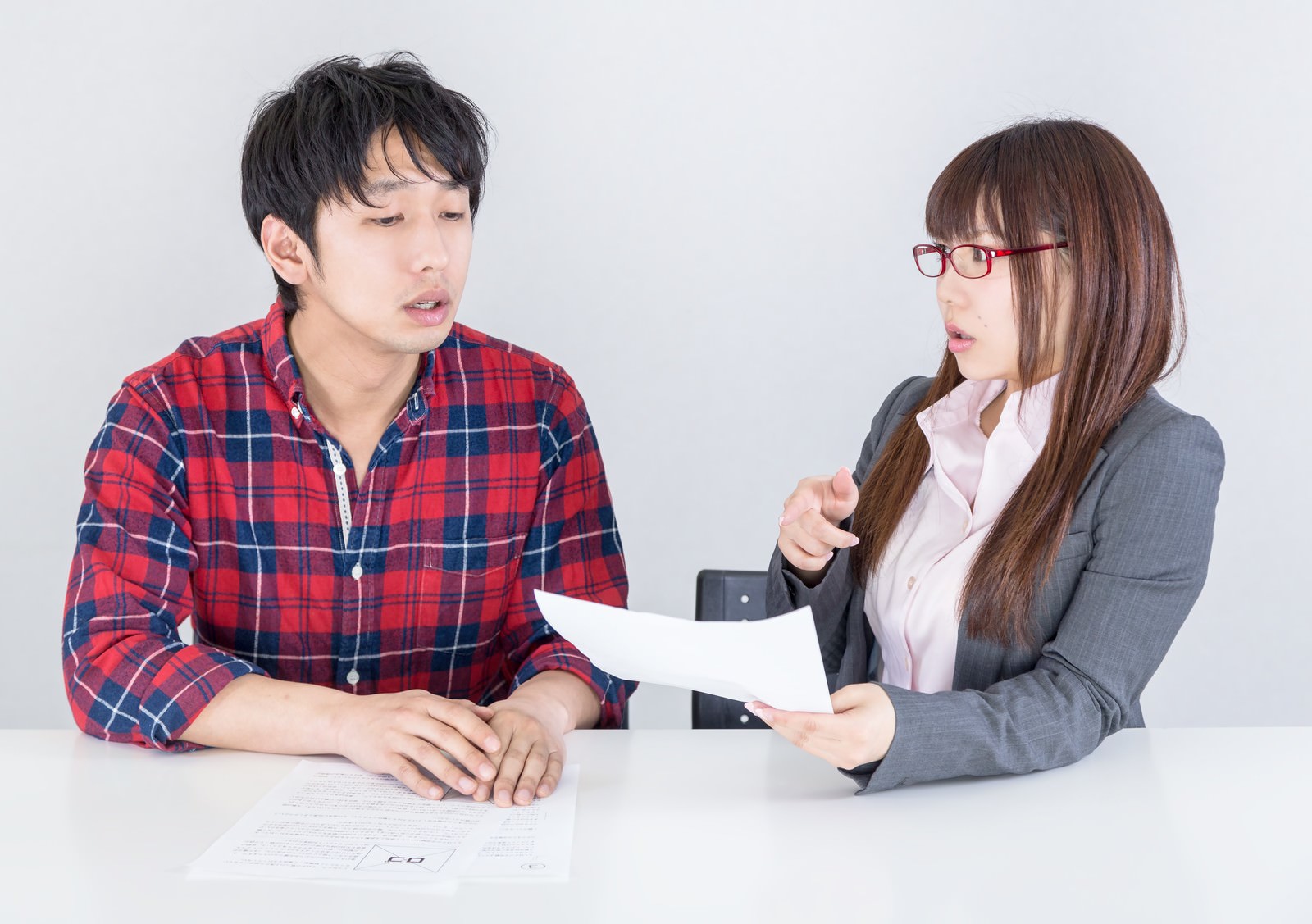 資料を指しながら説明する眼鏡の女性と、戸惑った表情で話を聞く男性の2人が向かい合っている［モデル：大川竜弥 Lala］