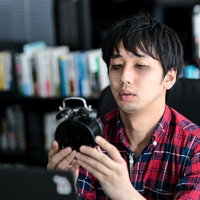 目覚まし時計を両手で持って見る赤チェックシャツの男性の写真