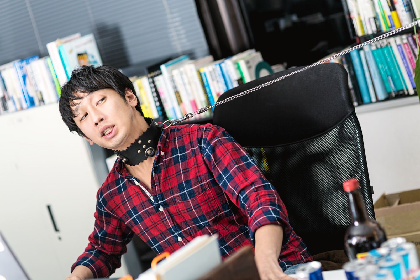 赤紺チェック柄シャツの男性が黒い首輪で拘束され、背後の本棚の前で上を向いて苦笑いしている［モデル：大川竜弥］