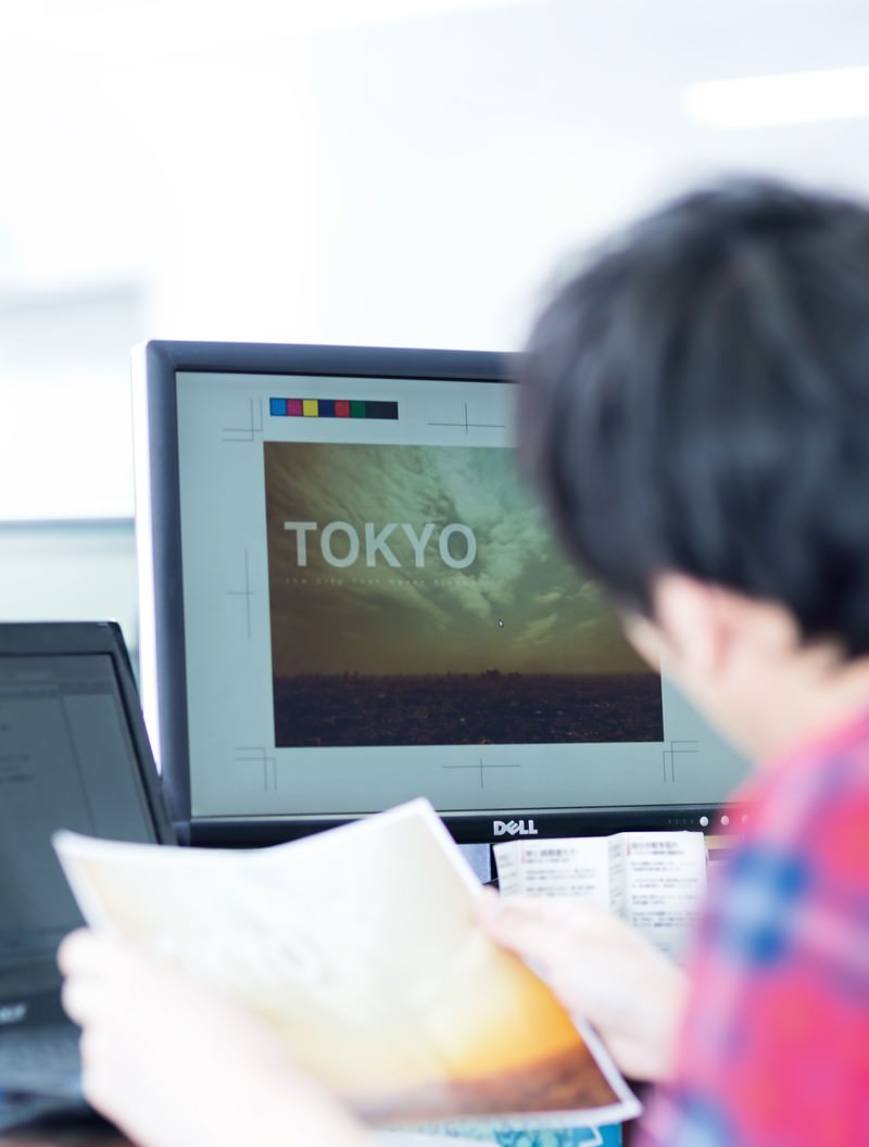 デザイン画面をモニターで確認するデザイナー男性が、手に持った資料を見ながら作業中［モデル：大川竜弥］