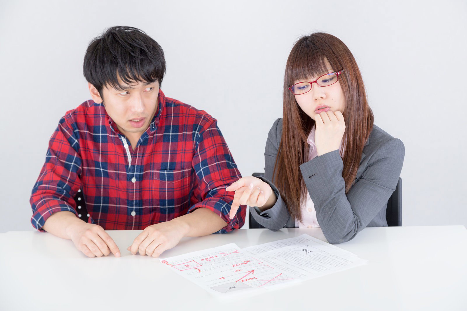 書類の修正指示を前に、指さす男性と考える表情の眼鏡をかけた女性［モデル：大川竜弥 Lala］