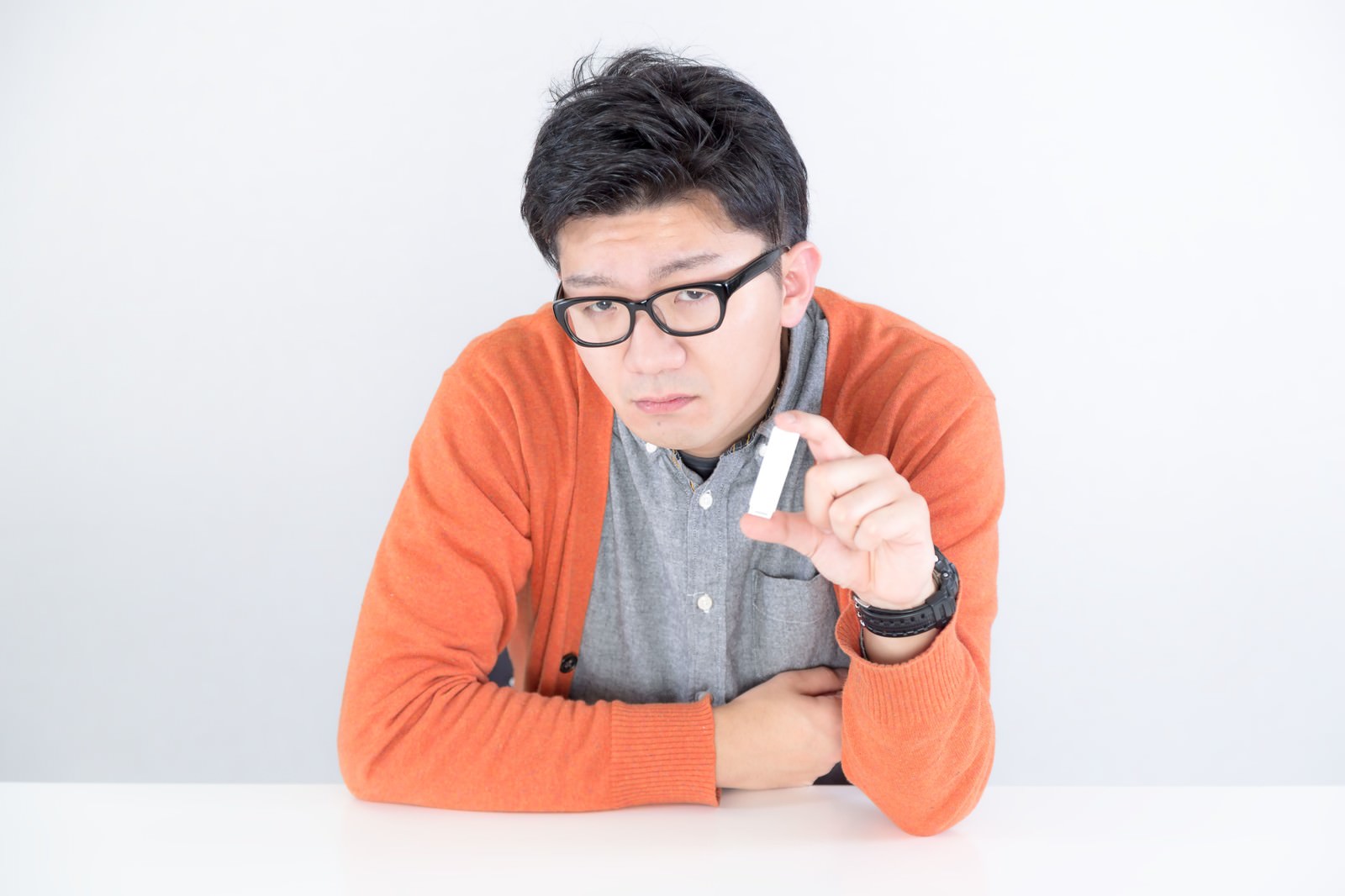 オレンジのカーディガンとグレーシャツを着た男性が眼鏡をかけ、机に腕をついてUSBメモリを手に持っている［モデル：OZPA］