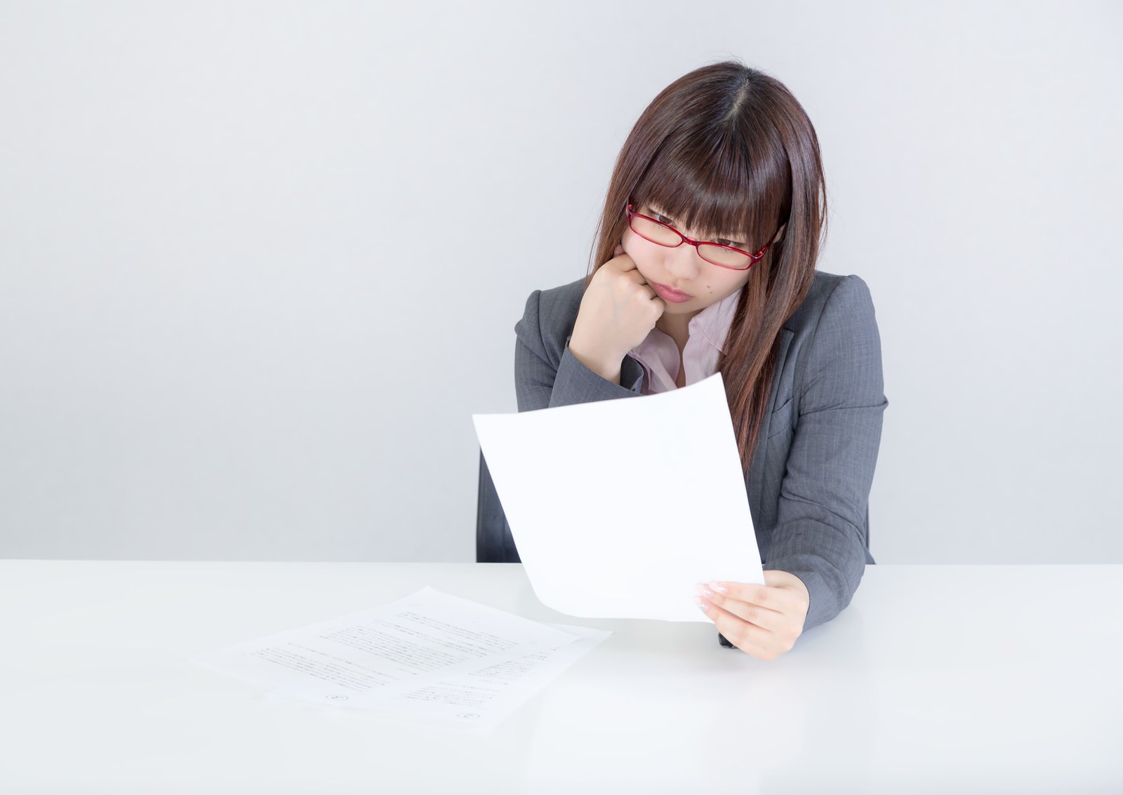 グレースーツの眼鏡をかけた女性がデスクで書類を持ち、真摯な表情で内容を確認している［モデル：Lala］