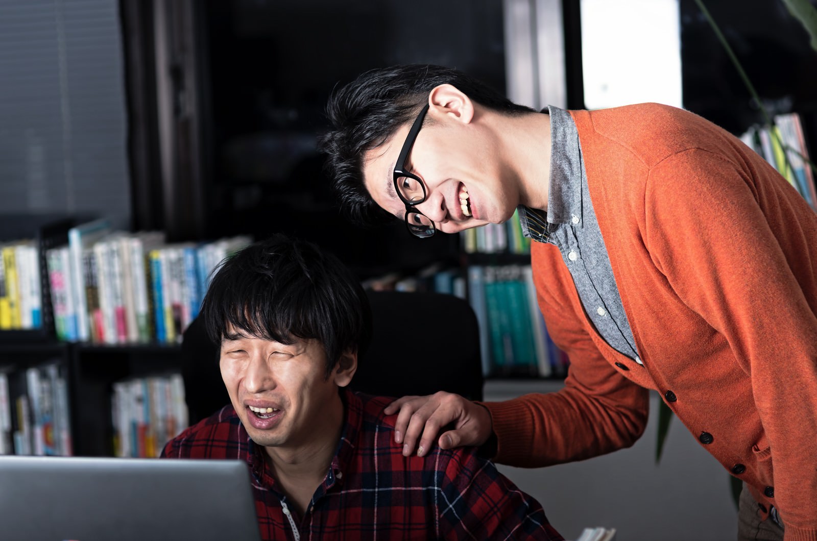 パソコンの前でうなだれるオレンジ色カーディガンの眼鏡男性、背景に本棚［モデル：大川竜弥 OZPA］