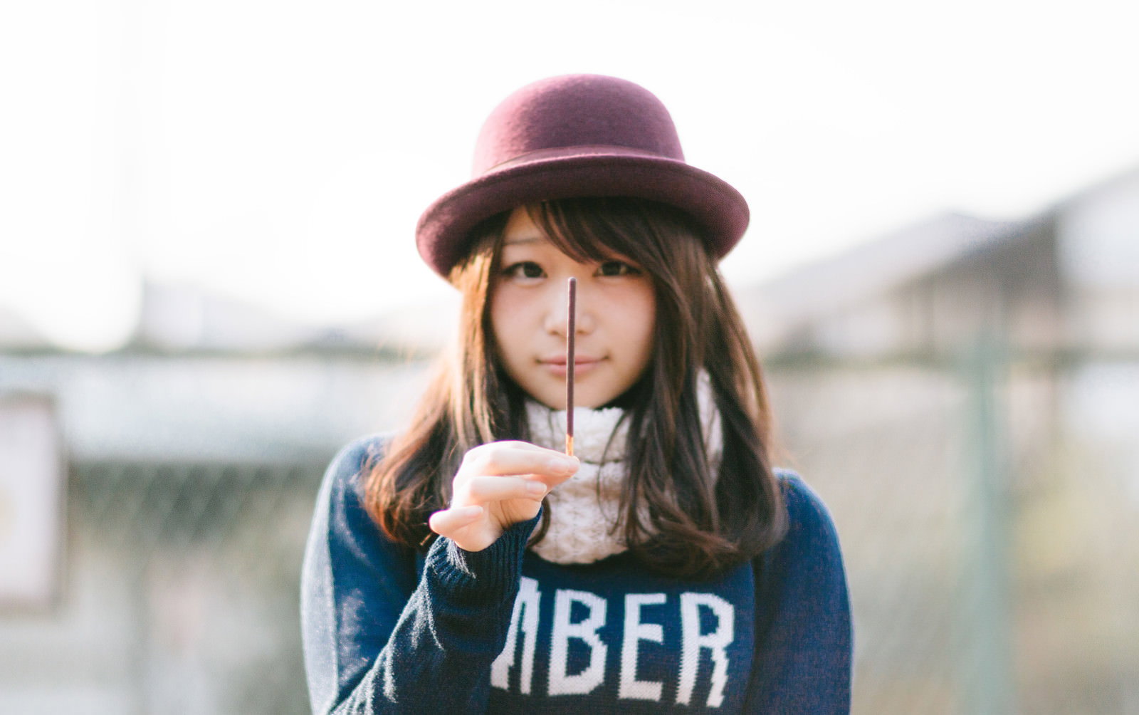 ピンク帽子とボーラーハット、紺トレーナー姿でポッキーをくわえほほえむ女性が河川沿いに立つ［モデル：茜さや］