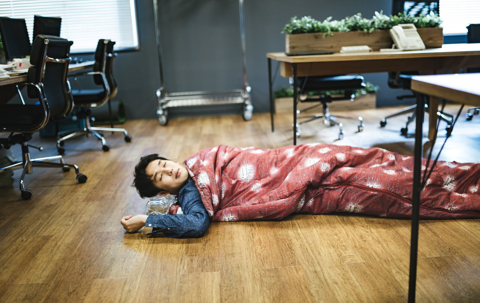 オフィスの床に赤白い寝袋を敷いて横たわり眠る男性エンジニア、背景に机と黒いチェア［モデル：大川竜弥］