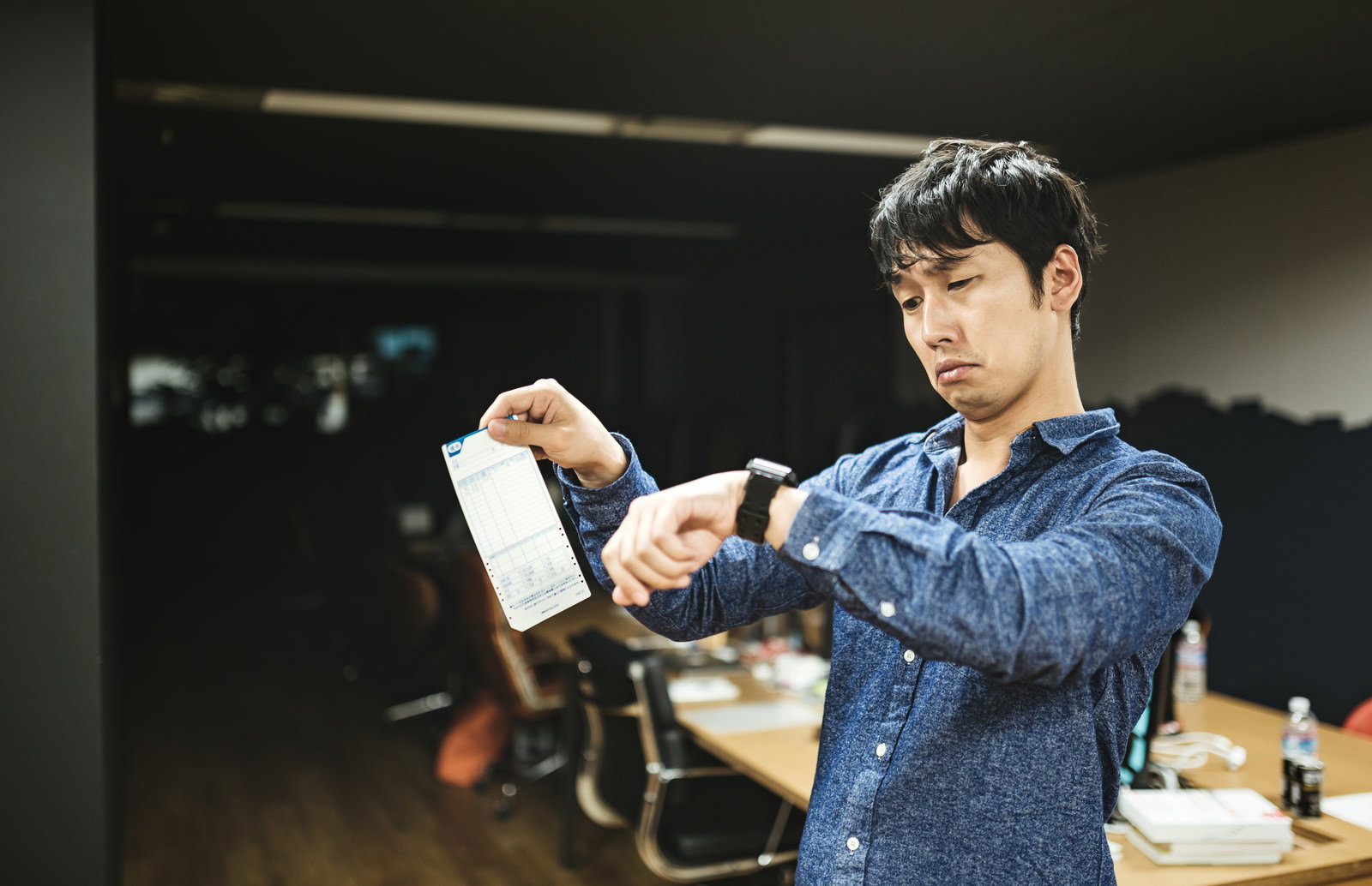 暗いオフィスでタイムカードと腕時計を確認する、疲労を滲ませた男性社員［モデル：大川竜弥］