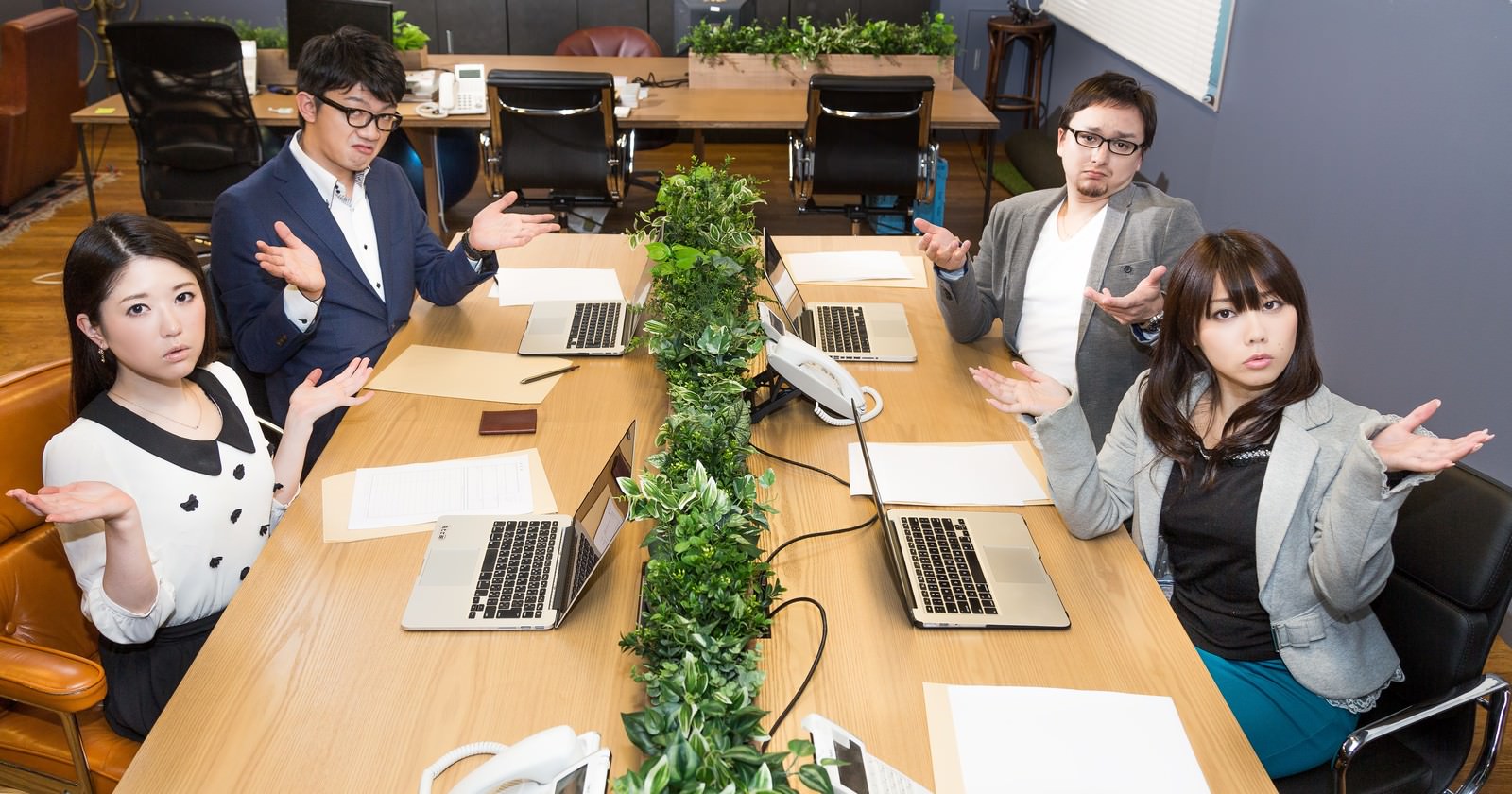 ノートPCと観葉植物がある会議テーブルで手を広げる4人［モデル：Max_Ezaki OZPA Lala シマヅ］