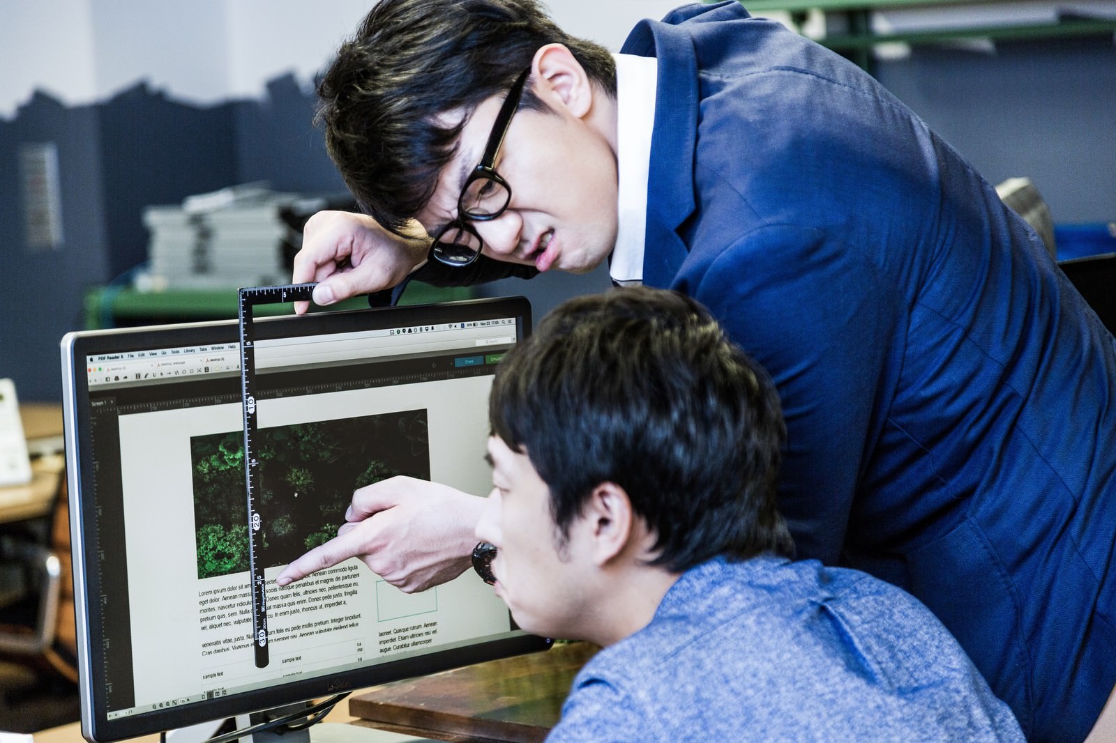 パソコン画面にディスプレイで定規を当てる青スーツの眼鏡男性［モデル：大川竜弥 OZPA］