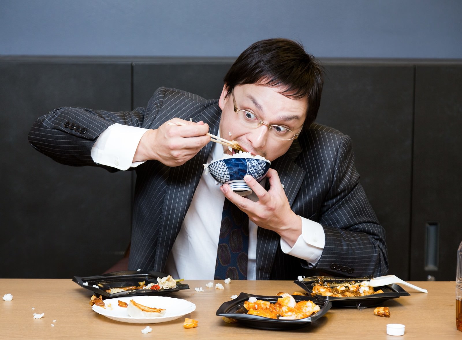 箸で食べ物を口に運ぶスーツの男性と黒い容器が並ぶテーブル［モデル：Max_Ezaki］