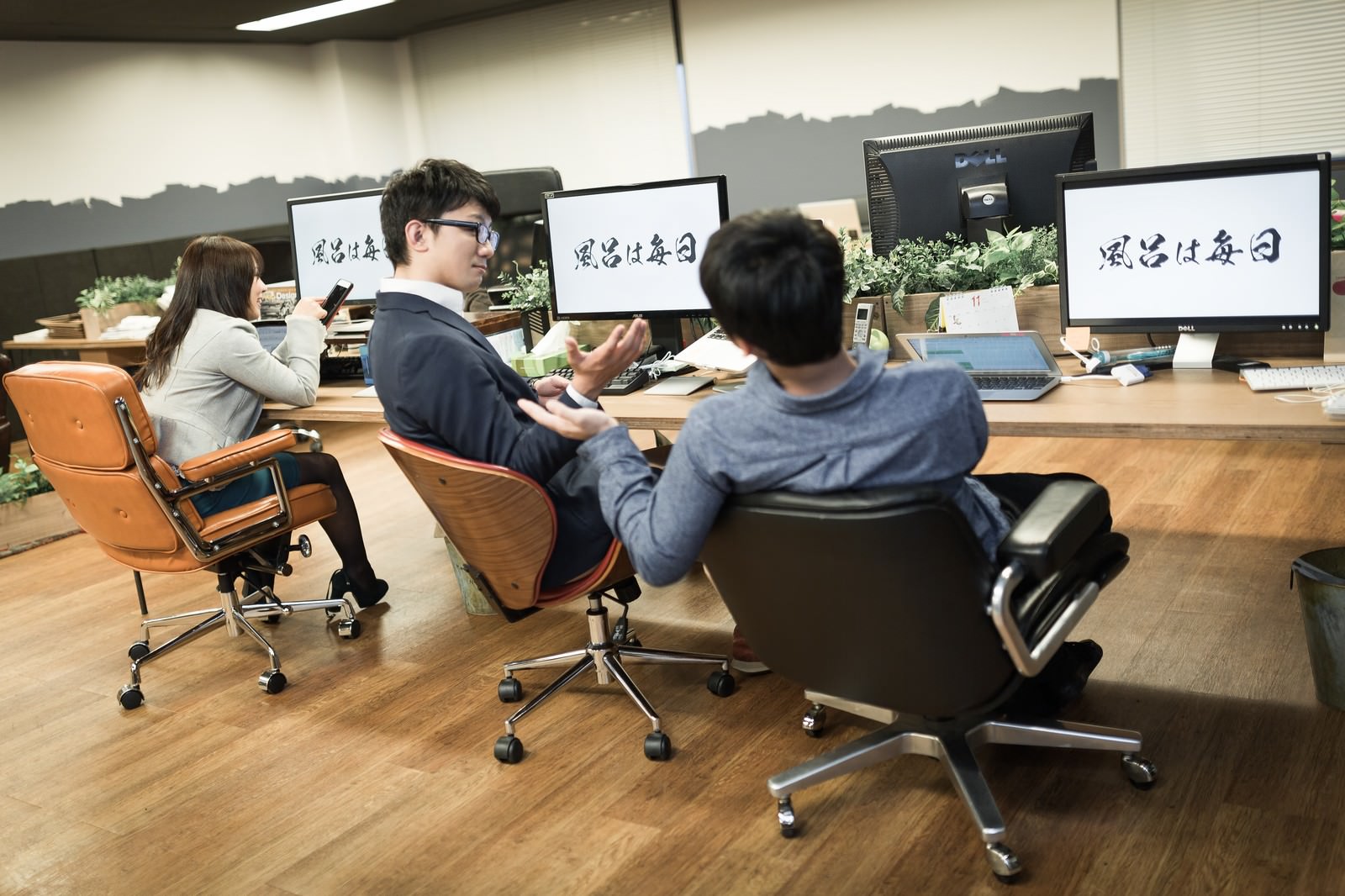 社訓が表示されたモニターの前で話す社員3人［モデル：大川竜弥 Lala OZPA］