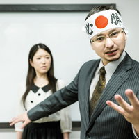 額に心と赤丸を書いた白い布を巻いて話すスーツ男性の写真