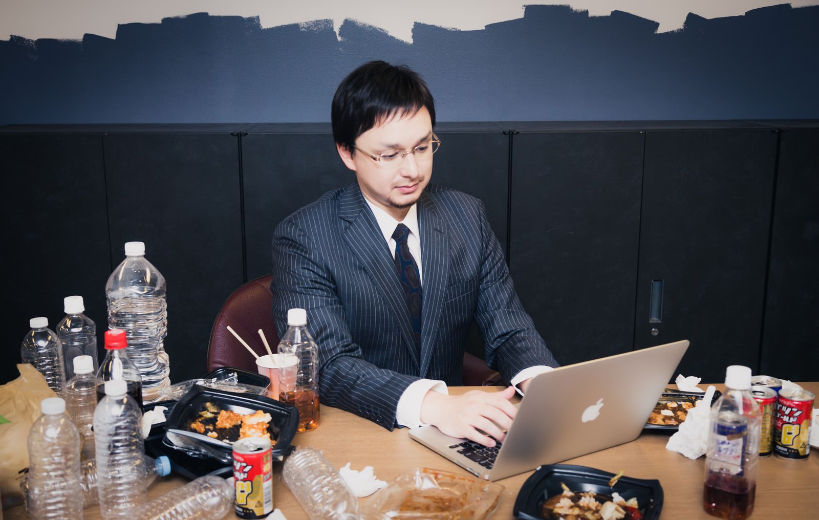 ペットボトルやカップ麺が散乱するデスクでPCを操作する縦縞スーツの男性［モデル：Max_Ezaki］
