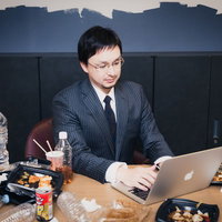 ペットボトルやカップ麺が散乱するデスクでPCを操作する縦縞スーツの男性の写真