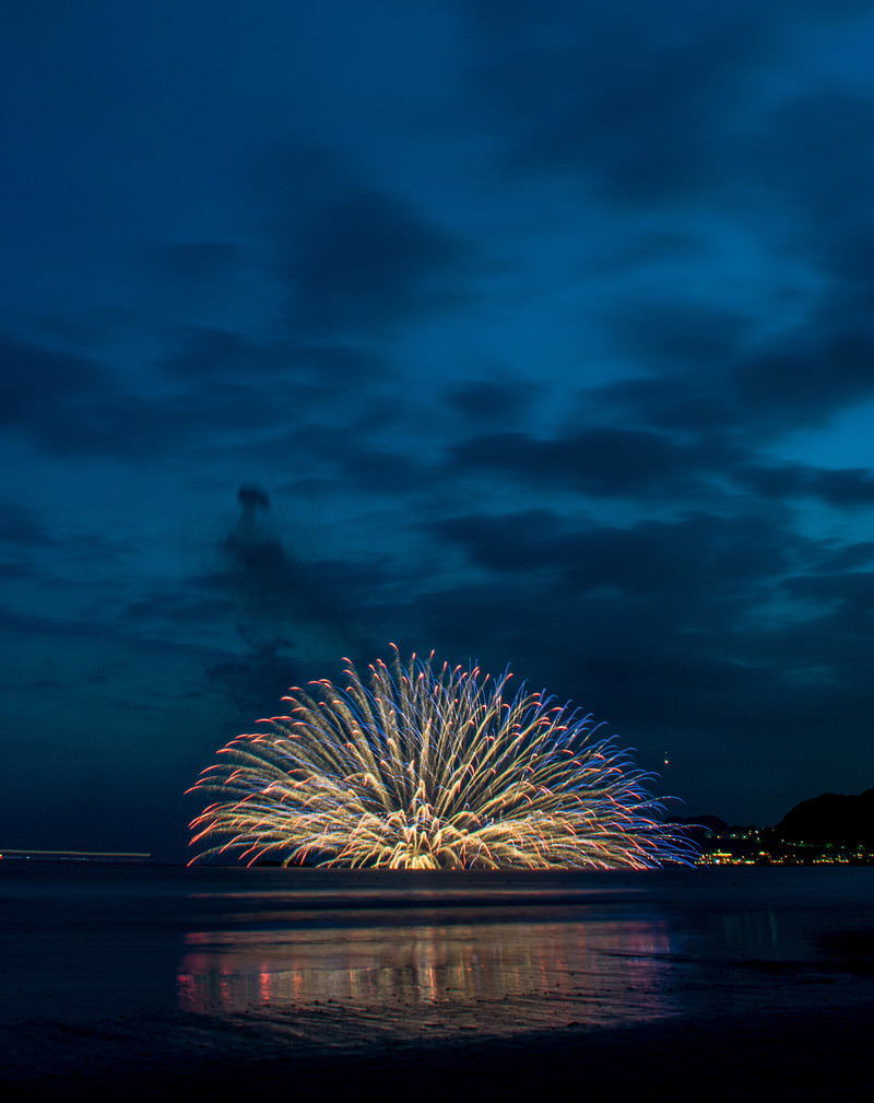 夜空に大きく開いた金色の水中花火と海面への光の反射