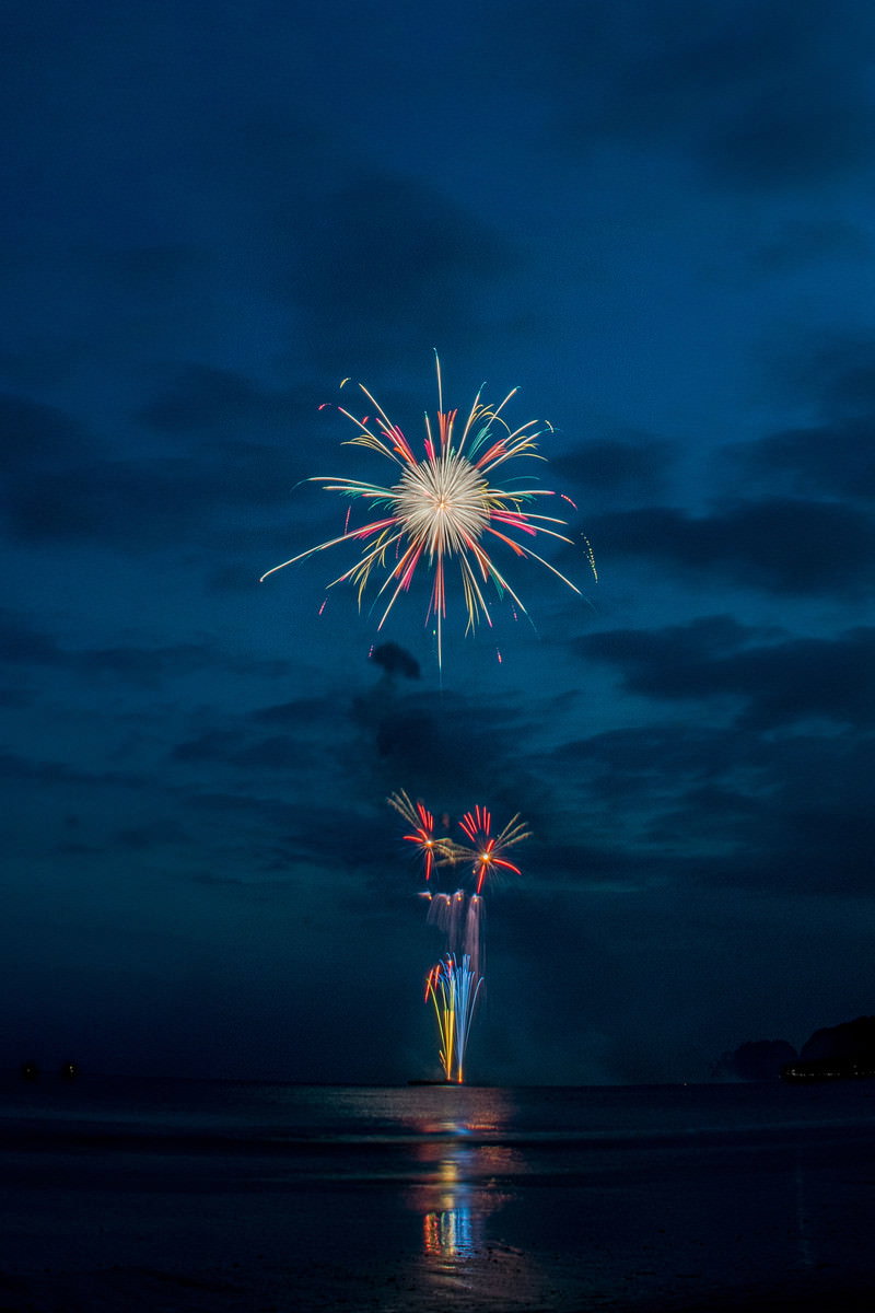 夜空に打ち上がった花火が海面に反射している砂浜からの風景
