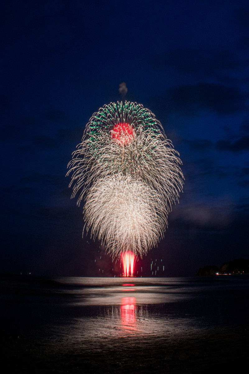 海上に打ち上がった大輪の白い花火が夜空に開き、海面に光が反射している様子