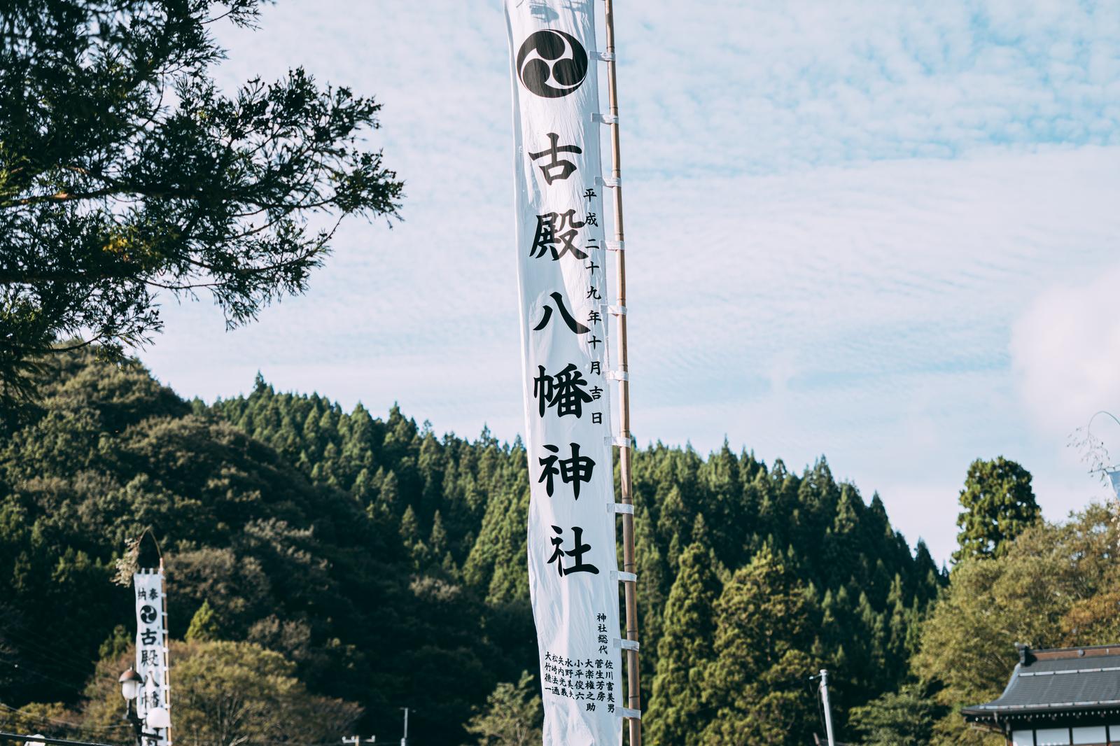 古殿八幡神社の白い幟に黒い文字で神社名と日付が書かれ、背景に杉林のある風景