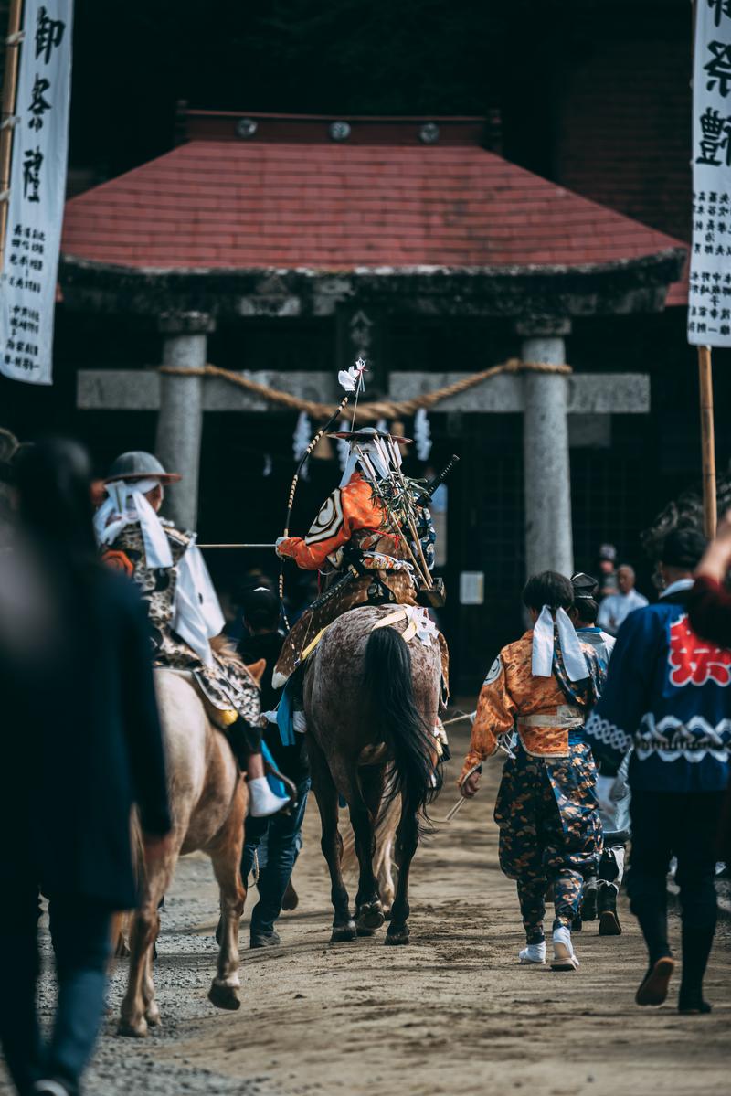 古殿八幡神社の境内で朱色の衣装を着た騎手が黒い馬に乗り、白い旗を持ちながら進んでいる