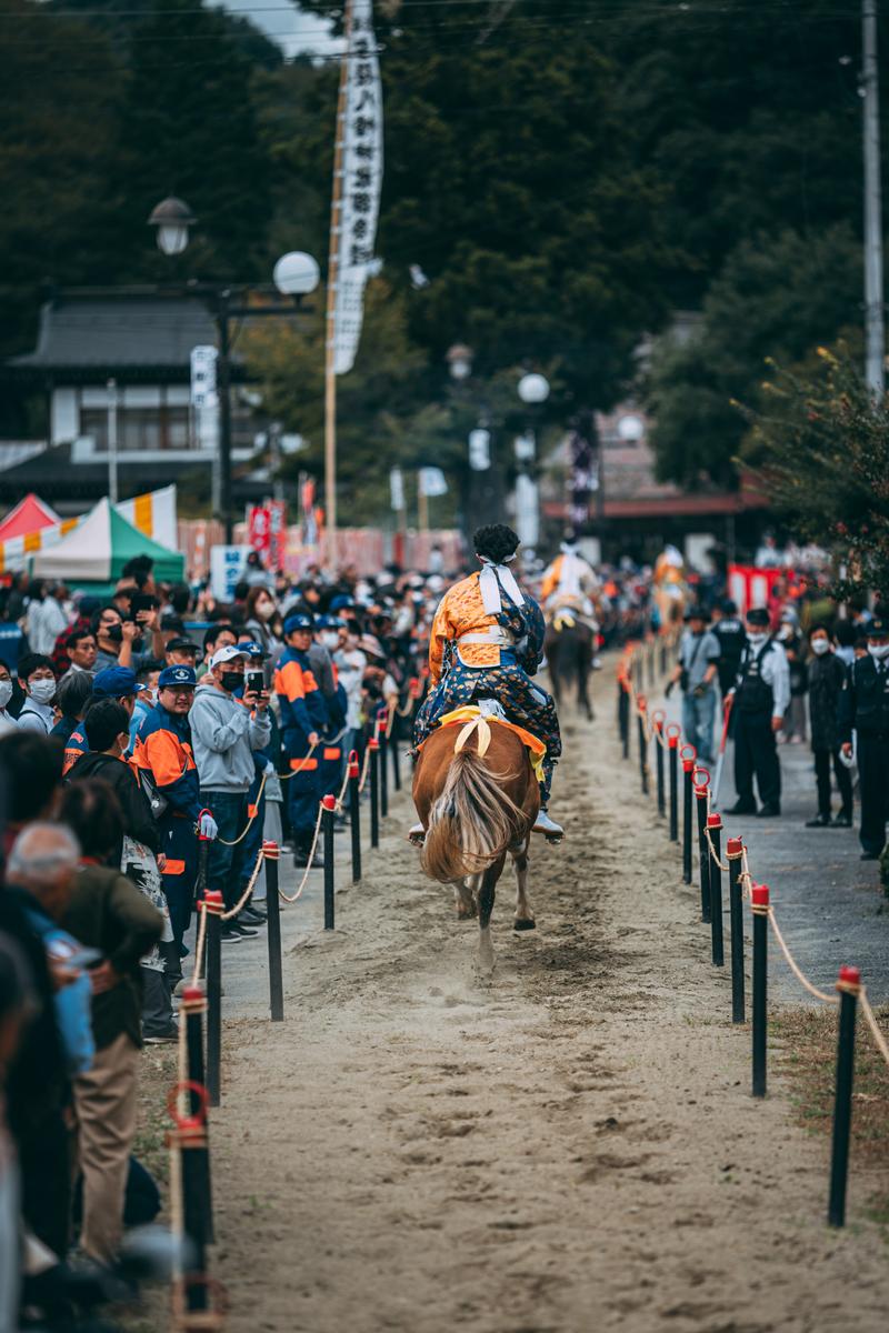 古殿八幡神社例大祭で、砂の参道を駆ける馬と装束を身につけた騎手、両側に立ち並ぶ観客たち