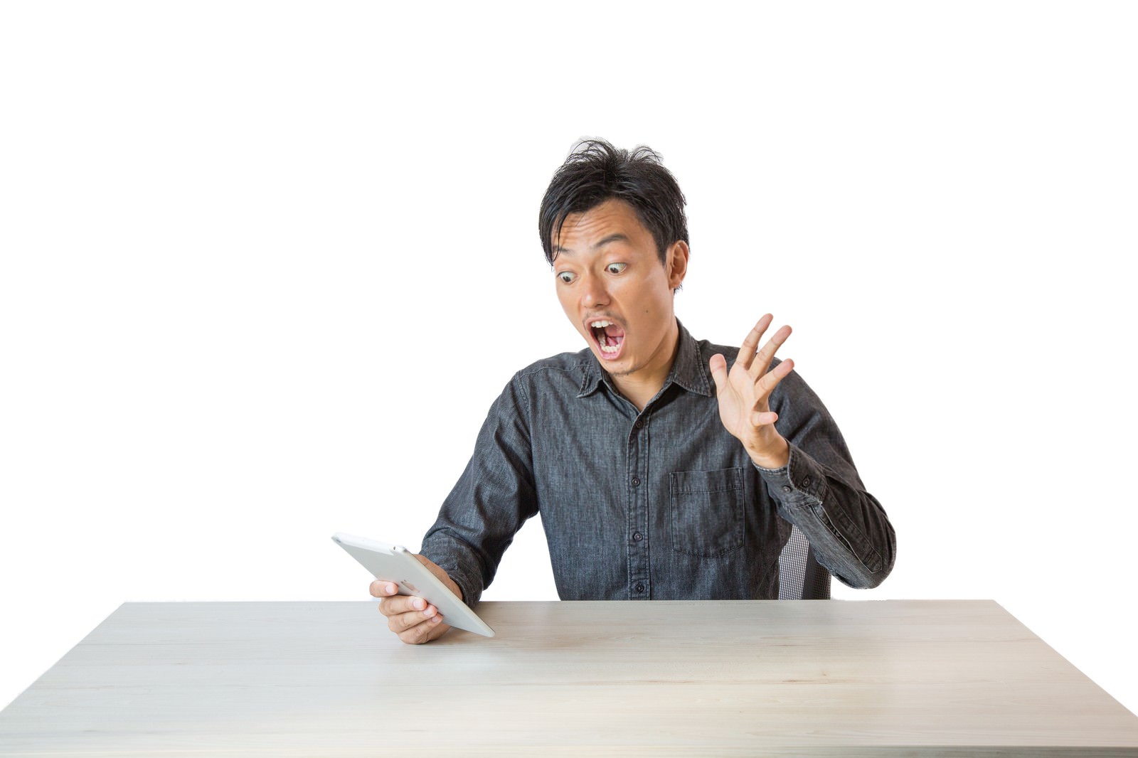 白背景のデスクでタブレットを持ちながら驚いた表情で指差しジェスチャーをする男性［モデル：藤沢篤］