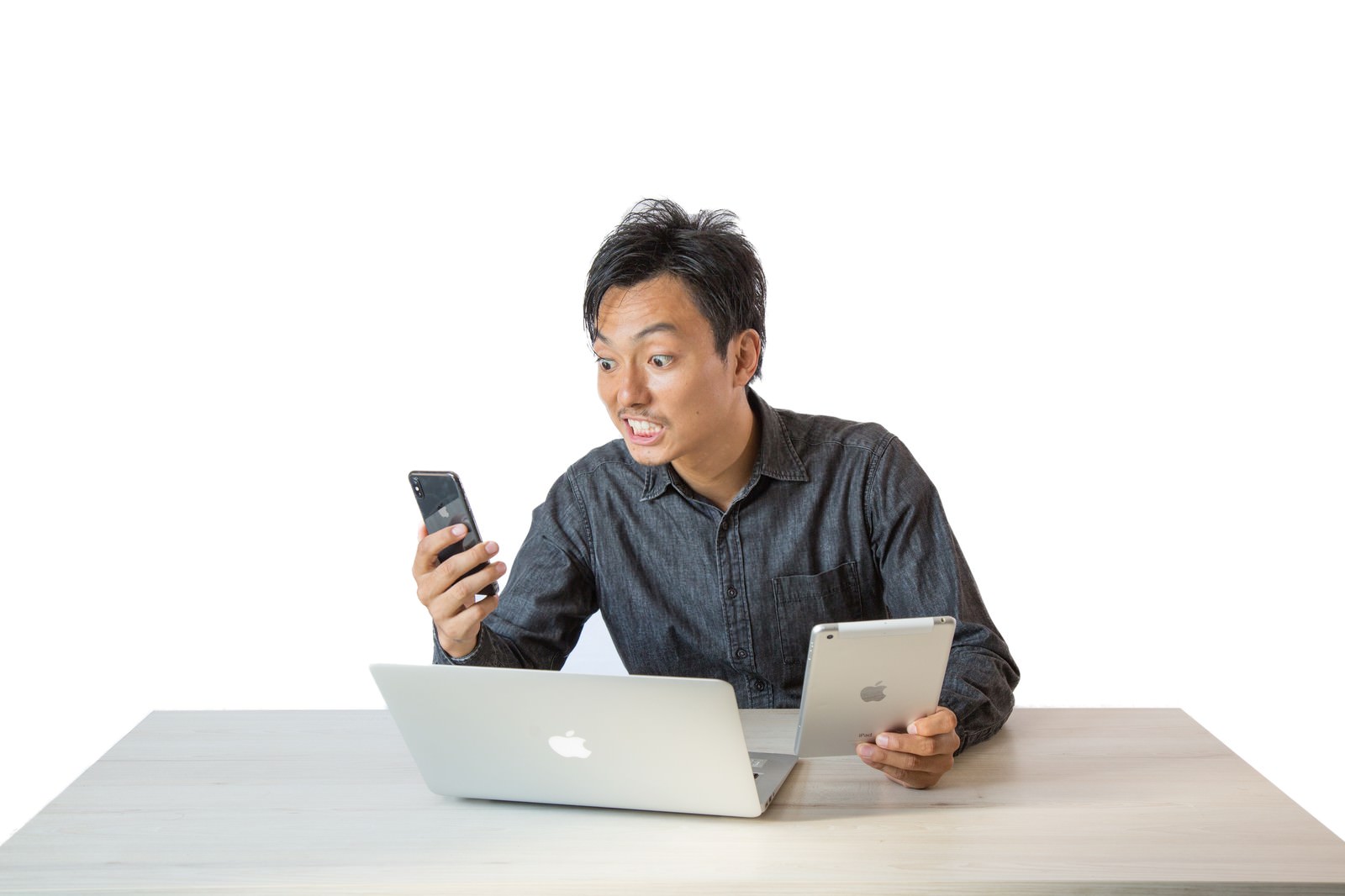 白い背景でノートパソコンの前に座り、スマートフォンを見て驚く男性［モデル：藤沢篤］