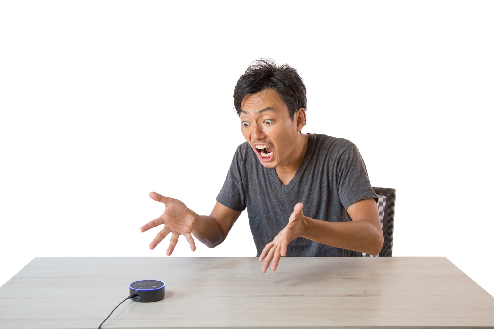 机の前で黒いスマートスピーカーに向かって両手を上げて困惑する男性［モデル：藤沢篤］