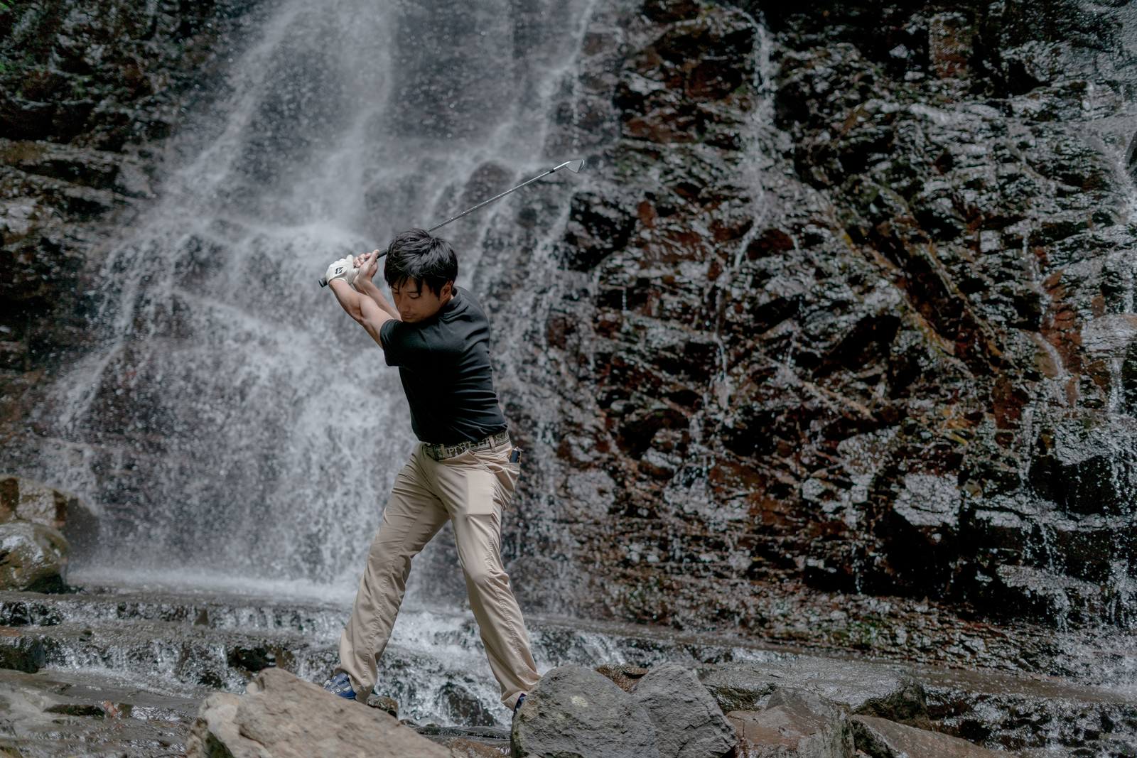 滝の水しぶきが飛び散る岩場でゴルフクラブを振る男性ゴルファー［モデル：サカモトリョウ］