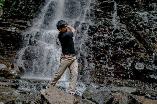 滝の岩場で難しいライからのアプローチショットを打つ男性ゴルファー