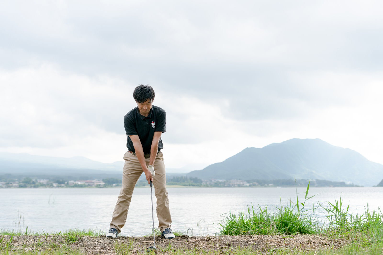 湖を背景にゴルフのアドレスの構えを取る男性ゴルファー［モデル：サカモトリョウ］
