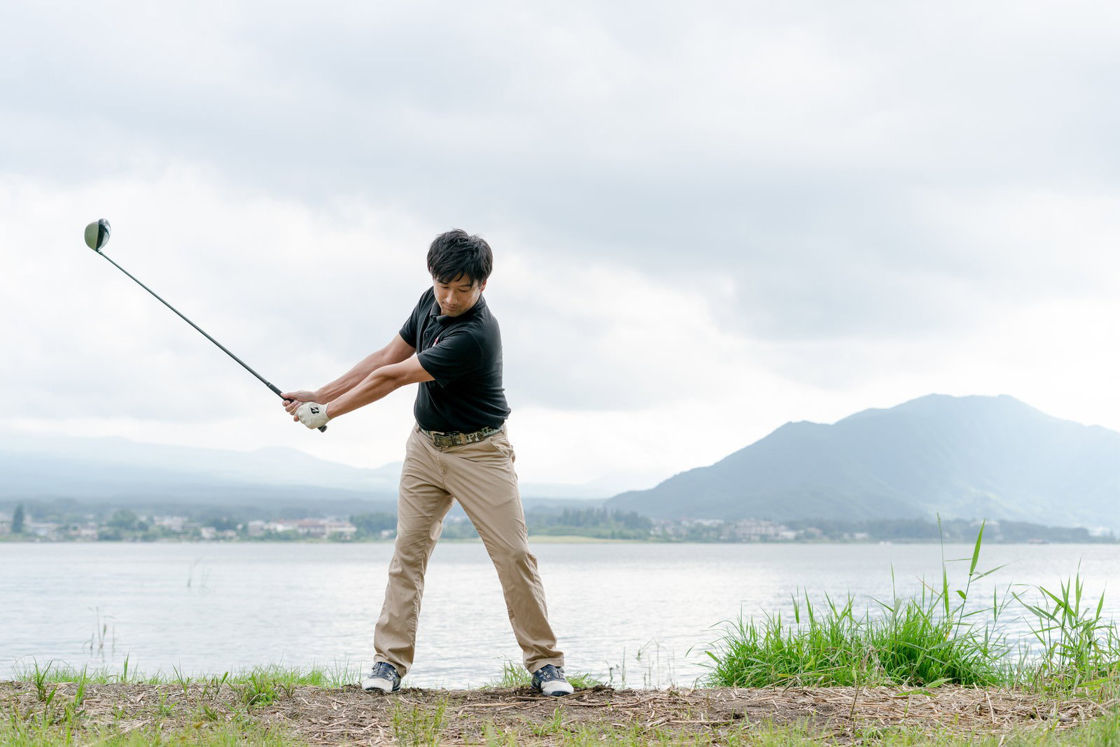 湖と山を背景にバックスイングをする男性ゴルファー［モデル：サカモトリョウ］