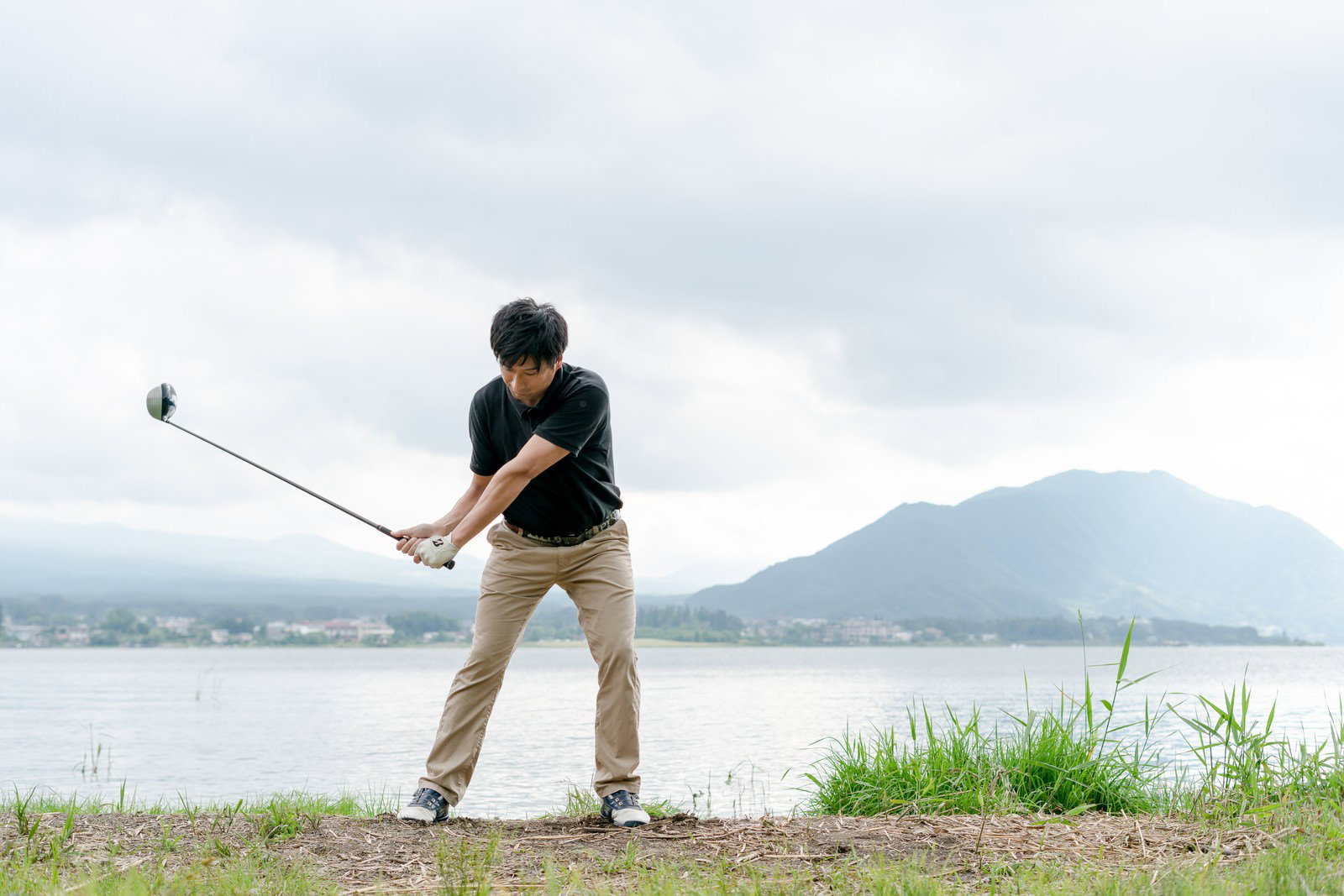 山と湖を背景にゴルフのバックスイングをする男性［モデル：サカモトリョウ］