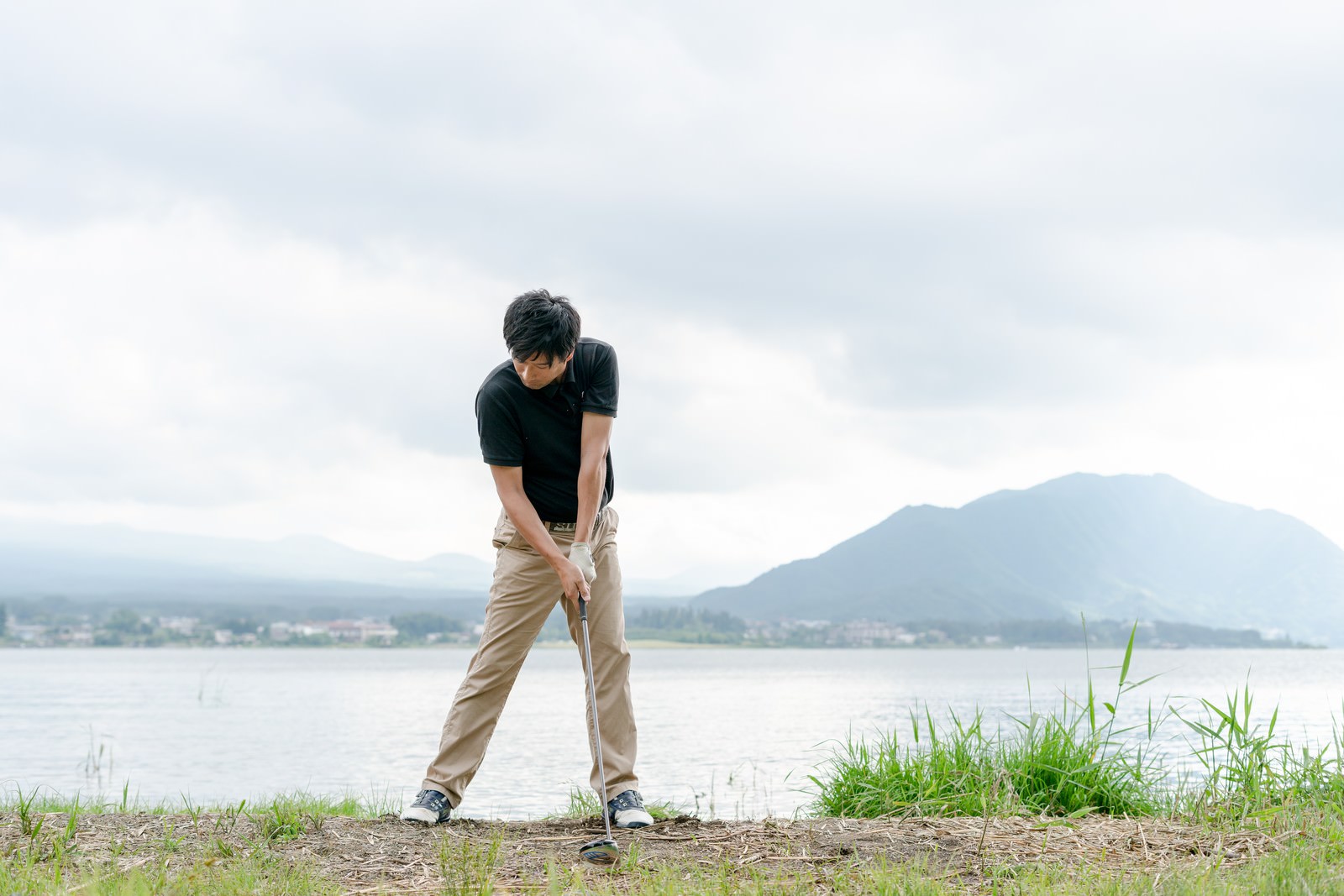 湖と山を背景にゴルフボールを打つ男性ゴルファー［モデル：サカモトリョウ］