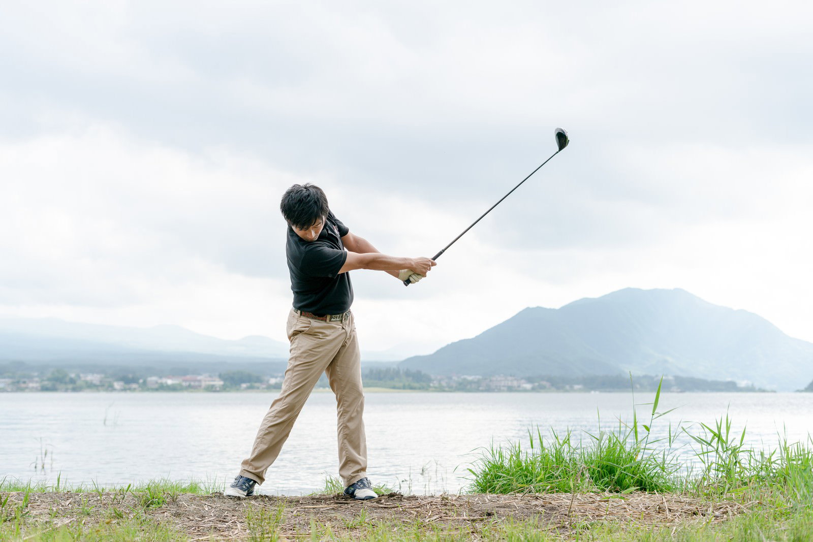 湖と山を背景にフォロースイングする男性ゴルファー［モデル：サカモトリョウ］