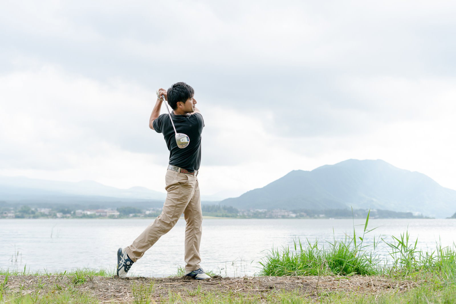湖と山を背景にドライバーでスイングする男性ゴルファー［モデル：サカモトリョウ］