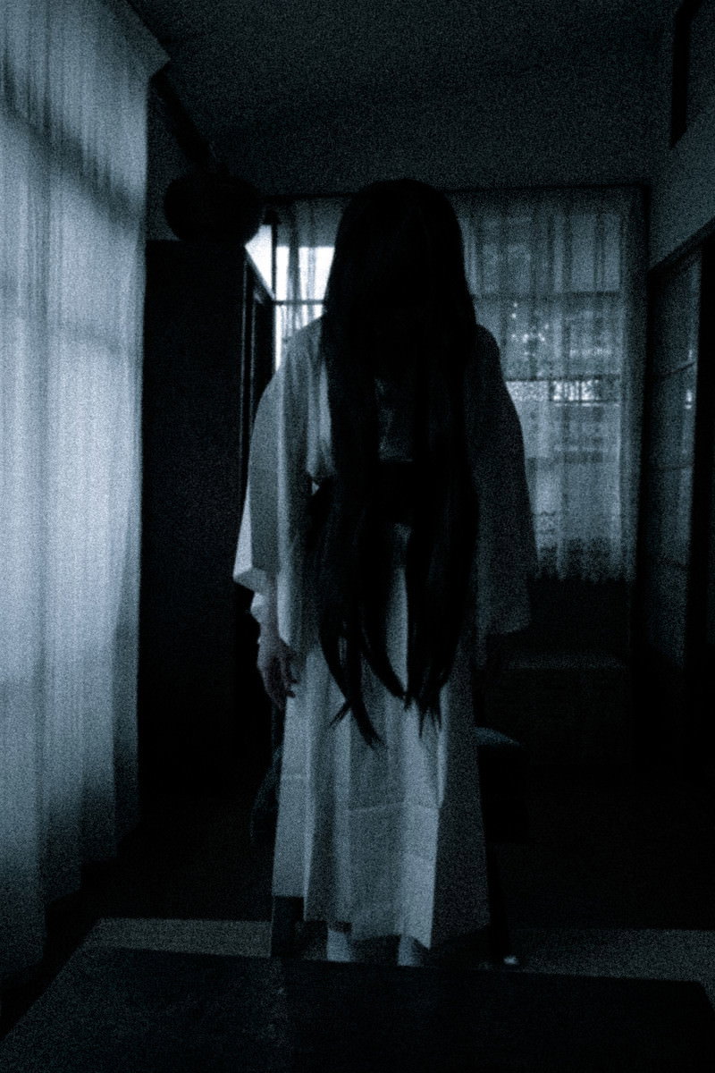 暗い室内で白い衣服を着た長髪の女性がうつむいて立っている［モデル：緋真煉］