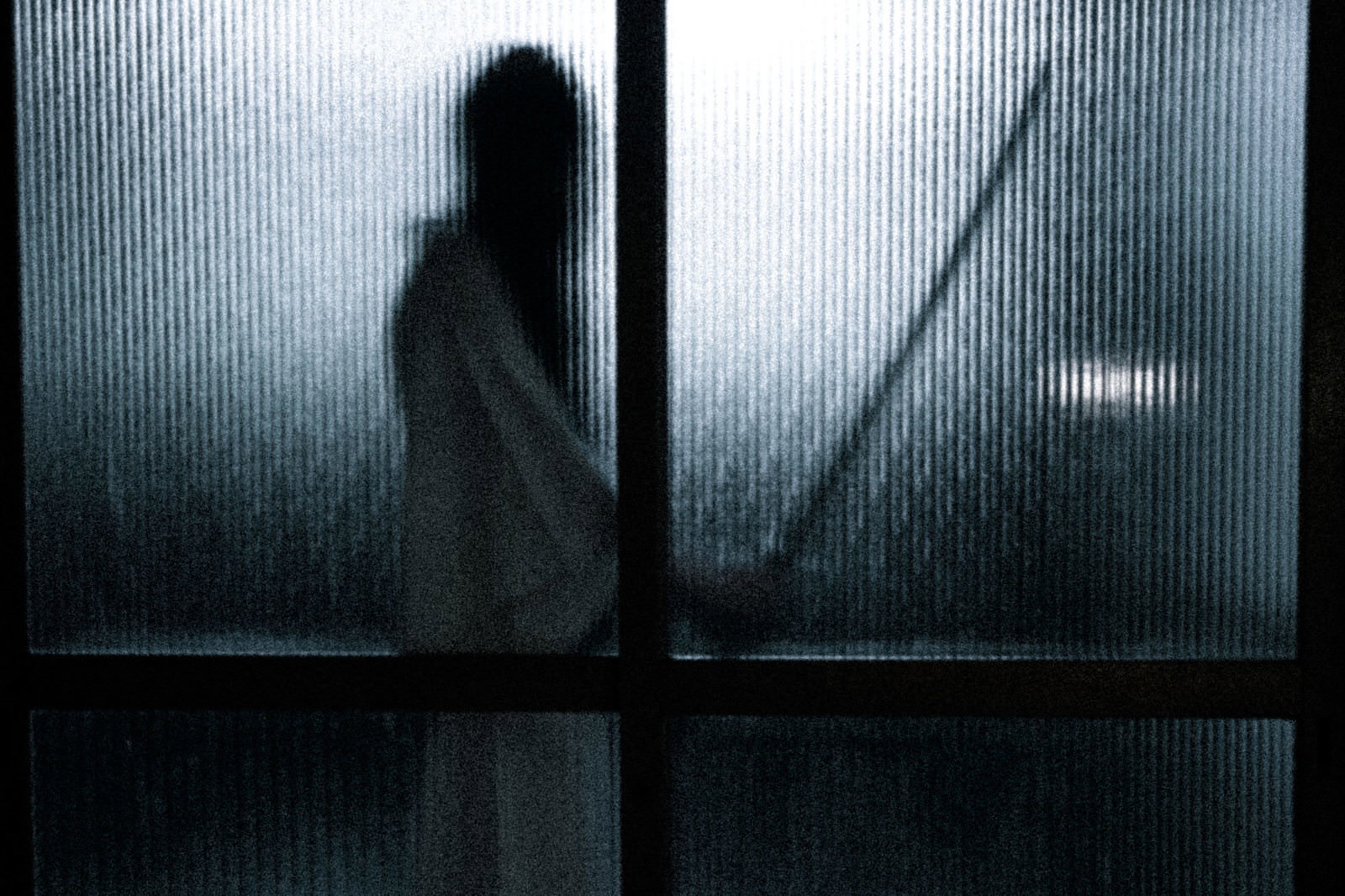 磨りガラス越しに刀を持った女性の人影が薄暗い空間に浮かび上がるホラーな光景［モデル：緋真煉］
