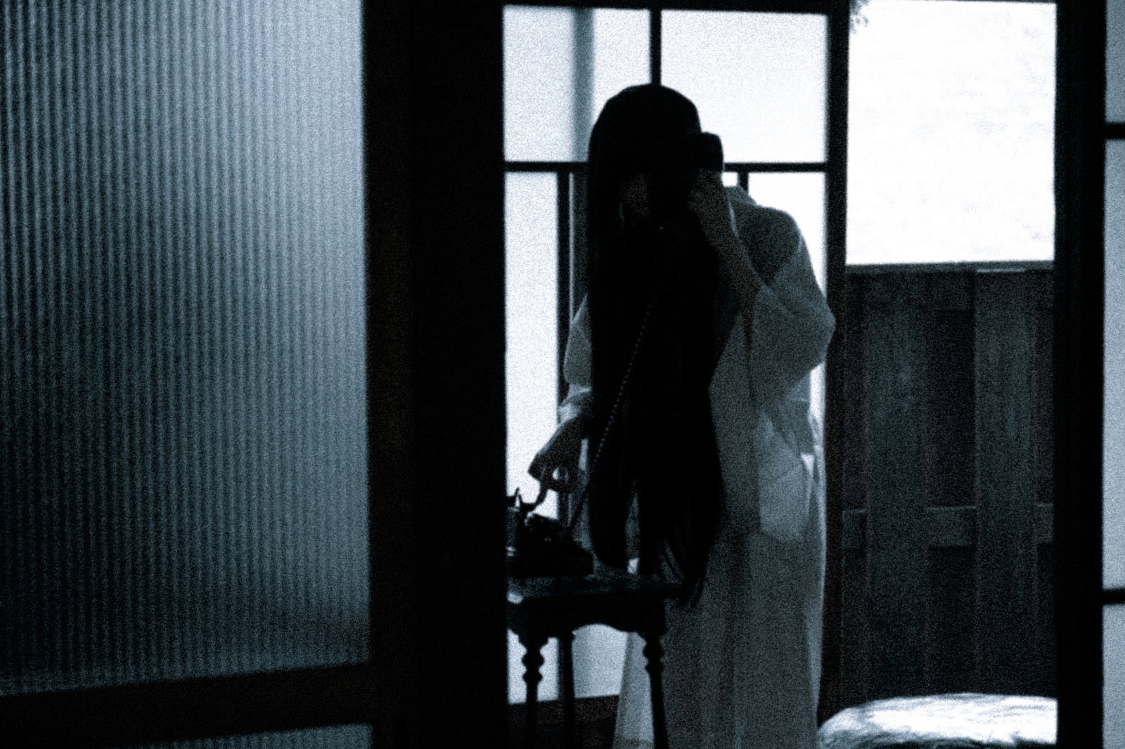 薄暗い室内で窓際に立ち黒電話をかける白い衣装の女性のシルエット［モデル：緋真煉］