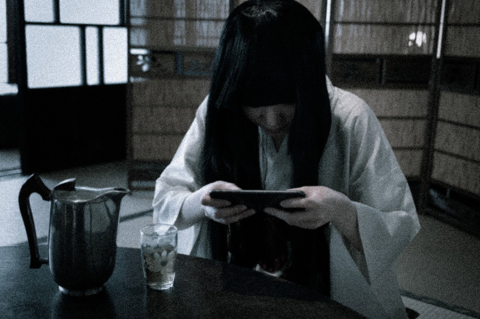 暗い部屋で白い服を着た長髪の女性がスマートフォンでゲームをしている［モデル：緋真煉］