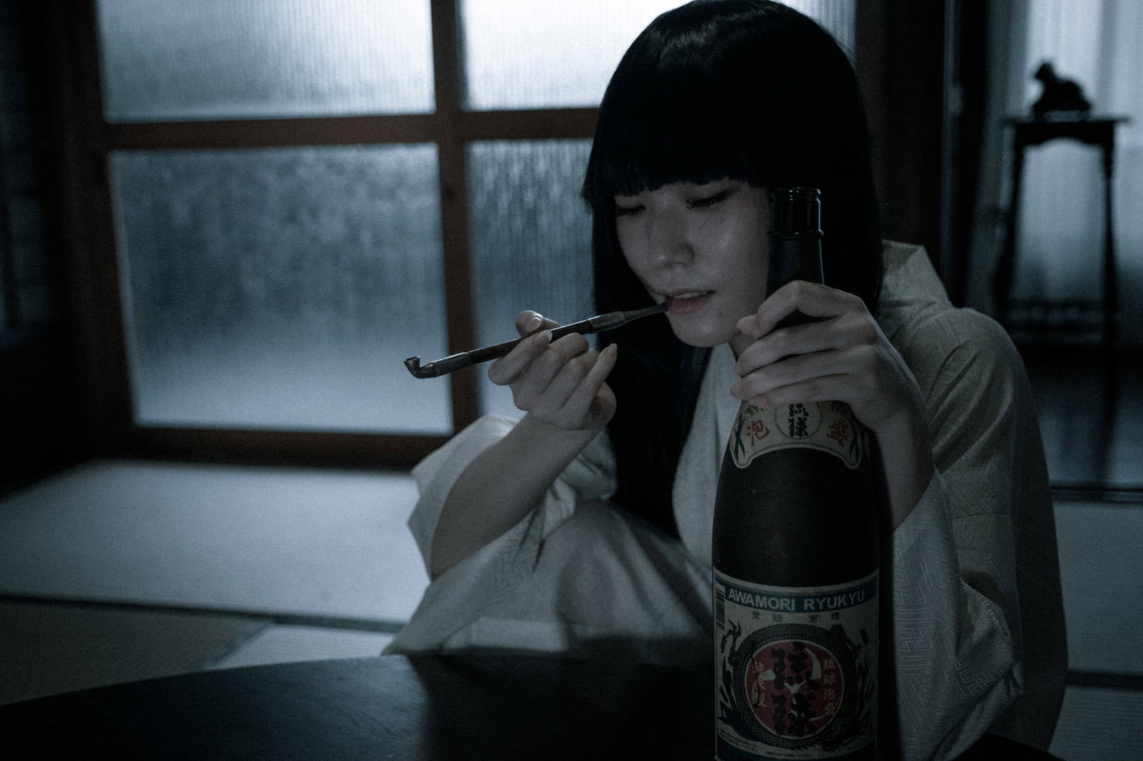 暗い室内でキセルを咥えて語りかける黒髪の女性と一升瓶［モデル：緋真煉］