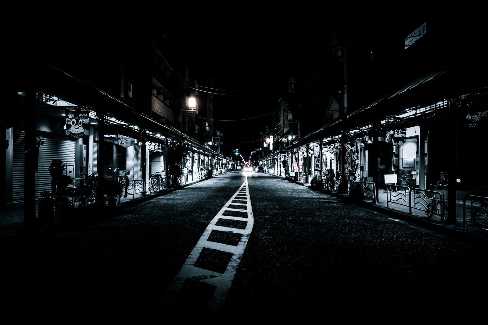 夜間に照明が灯る商店街。両側にシャッターが閉じた店舗が立ち並び、白い道路標線が奥へ伸びている